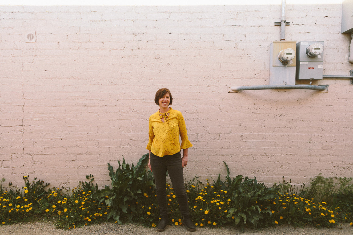 The designer wears a yellow shirt and stands outside in front of a light colored brick building lined with dandelions in bloom.
