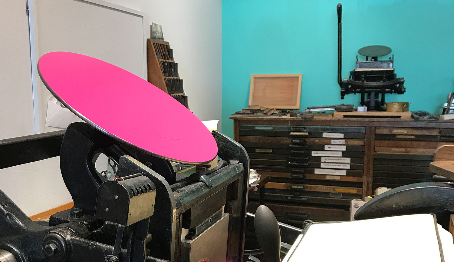 A vintage printing press in use with magenta pink ink is shown with printing equipment of the same era against a bright turquoise colored wall.
